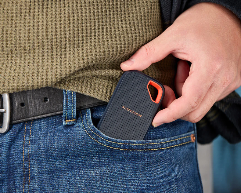 Osoba wkłada przenośny dysk SSD SanDisk do kieszeni niebieskich dżinsów, ubrana w zielony sweter.