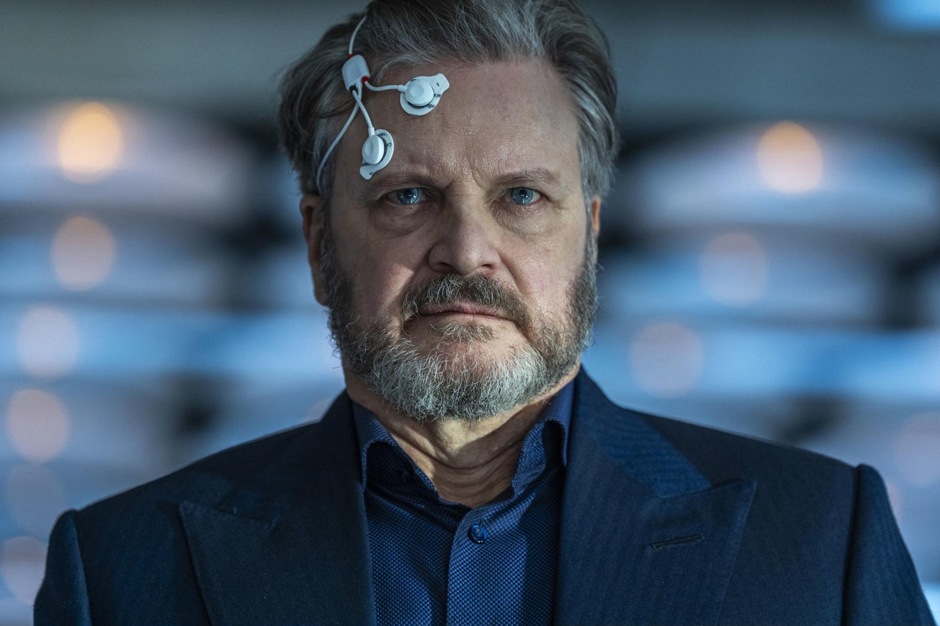 Ein Mann mit grauem Bart und blauen Augen, EEG-Sensoren an der Stirn und einem dunkelblauen Anzug.