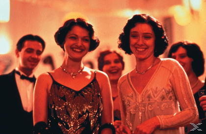 Zwei lächelnde Frauen in eleganter Abendkleidung stehen bei einer Feier im Mittelpunkt, umgeben von anderen Gästen.