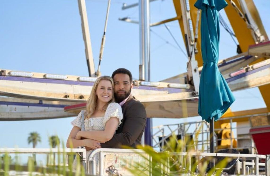 Ein glückliches Paar steht vor einem Riesenrad und einem blauen Regenschirm an einem sonnigen Tag.