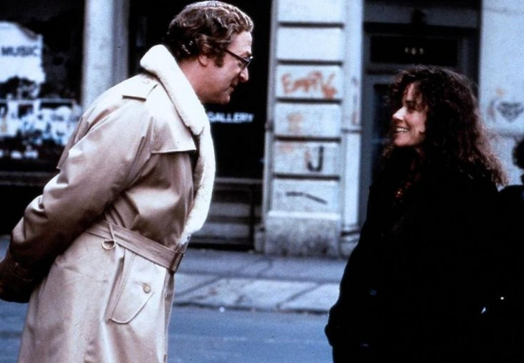 Ein Mann im hellen Trenchcoat mit Fellkragen unterhält sich auf der Straße mit einer lächelnden Frau.