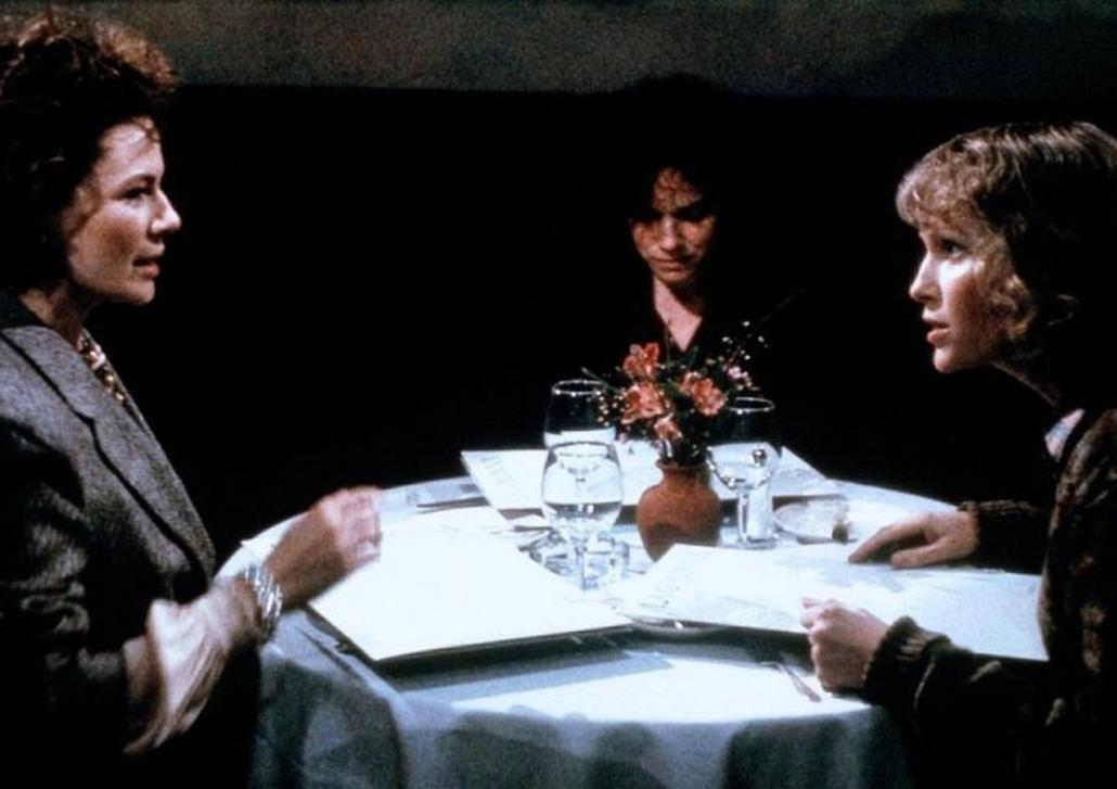 Zwei Frauen sitzen an einem runden Tisch in einem Restaurant und sprechen miteinander, während ein Kellner im Hintergrund steht.