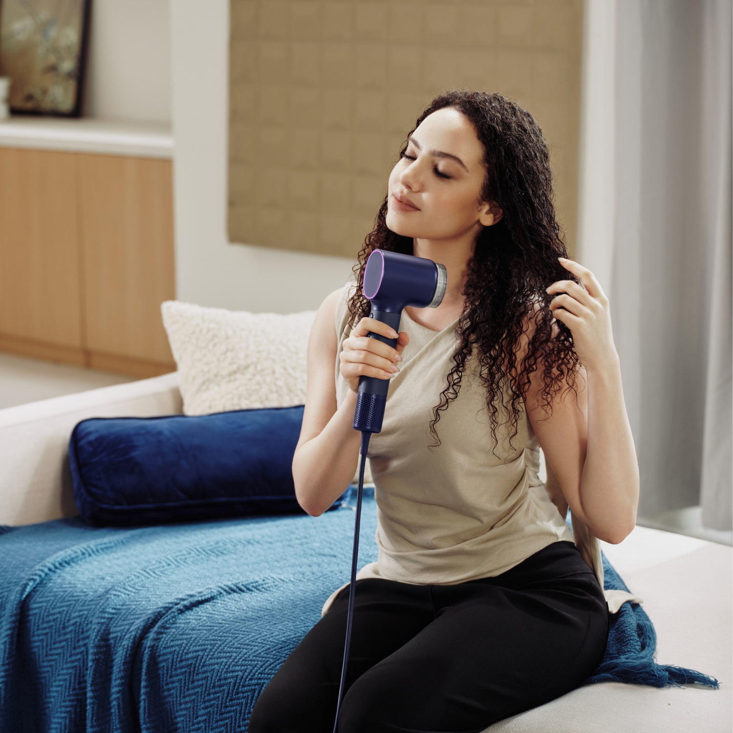 Une femme aux cheveux bouclés s'assoit sur un lit et sèche ses cheveux avec un sèche-cheveux bleu.