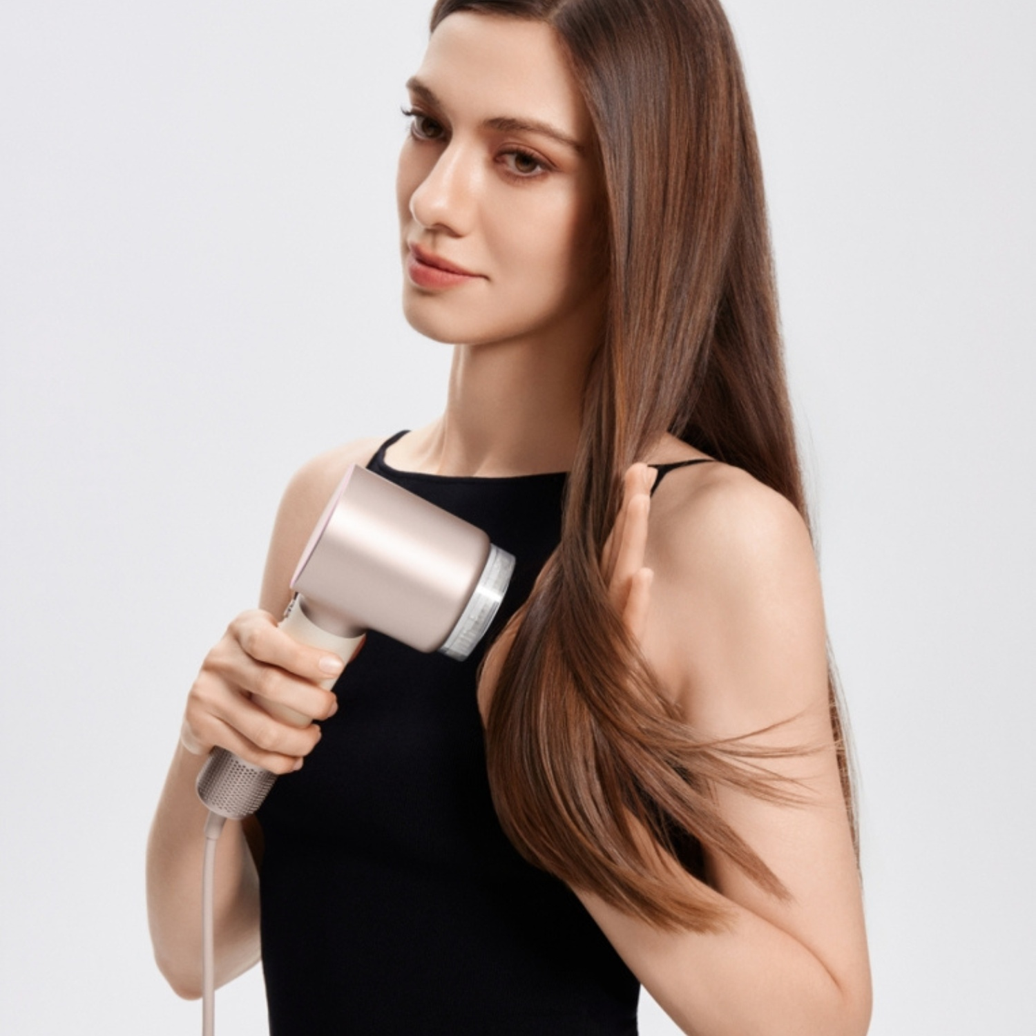 Une femme aux longs cheveux bruns sèche ses cheveux avec un sèche-cheveux or rose sur fond blanc.