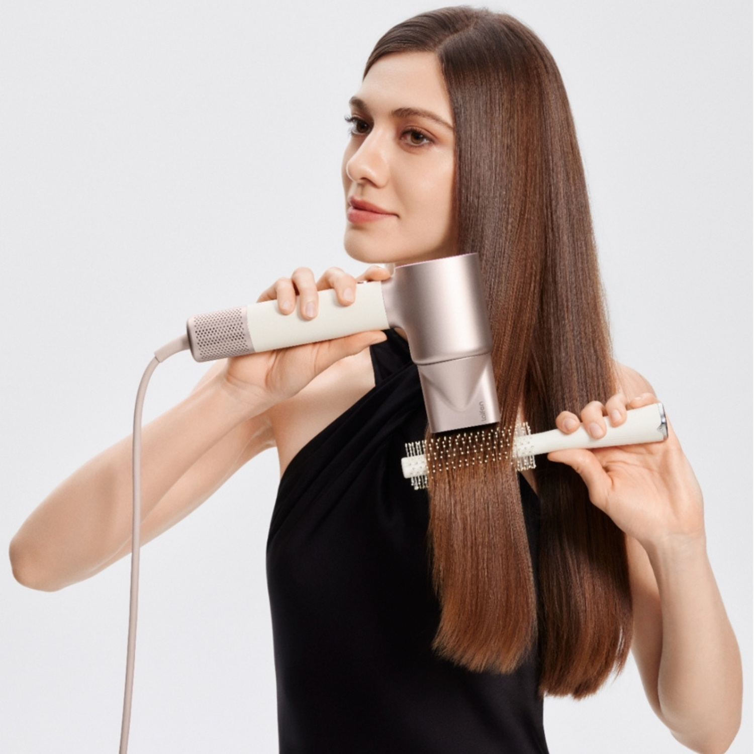 Une femme aux longs cheveux bruns se sèche et se brosse les cheveux avec un sèche-cheveux or rose et une brosse blanche.