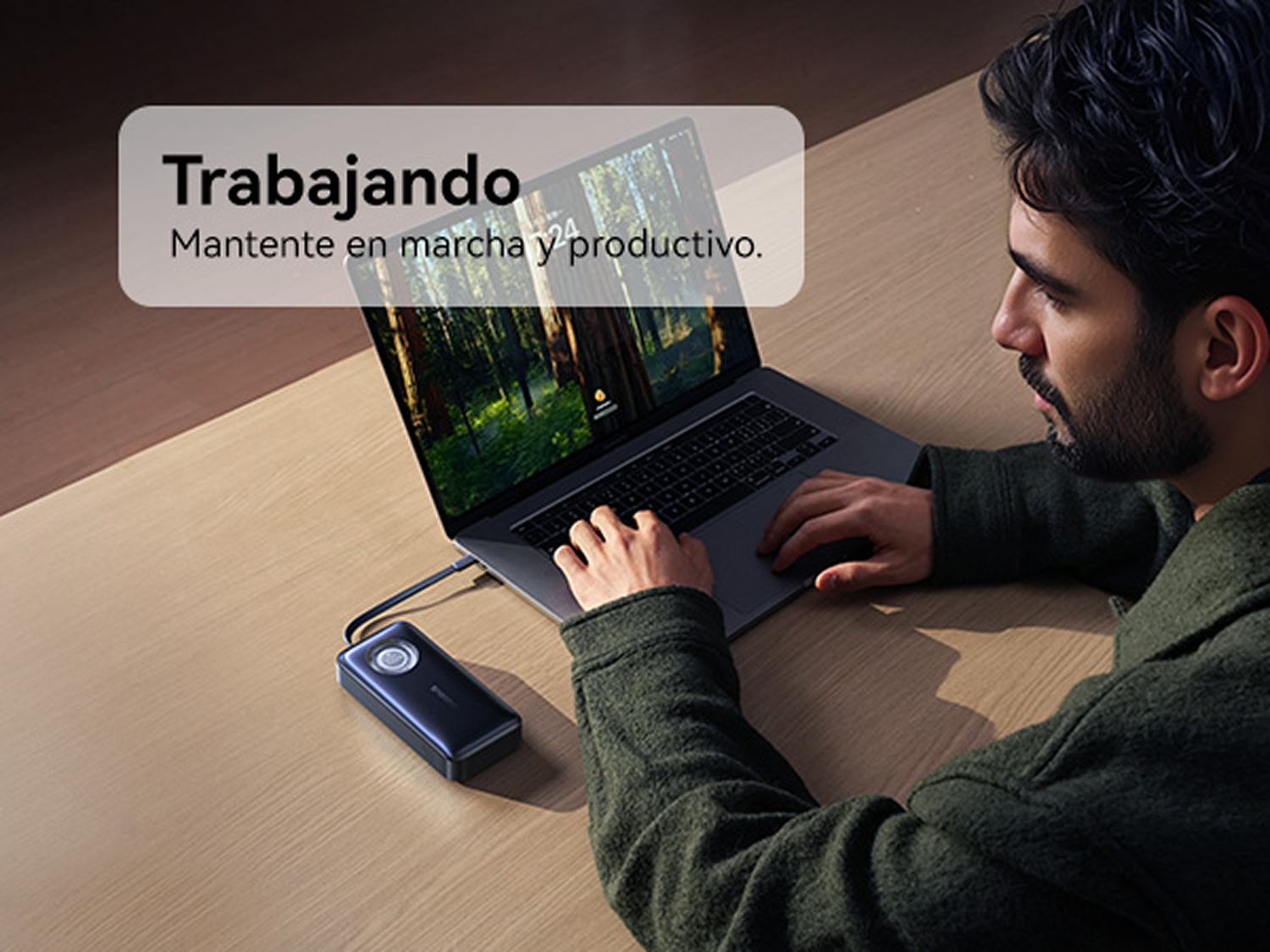 Un hombre con chaqueta verde escribe en una laptop conectada a una batería portátil en un escritorio de madera.