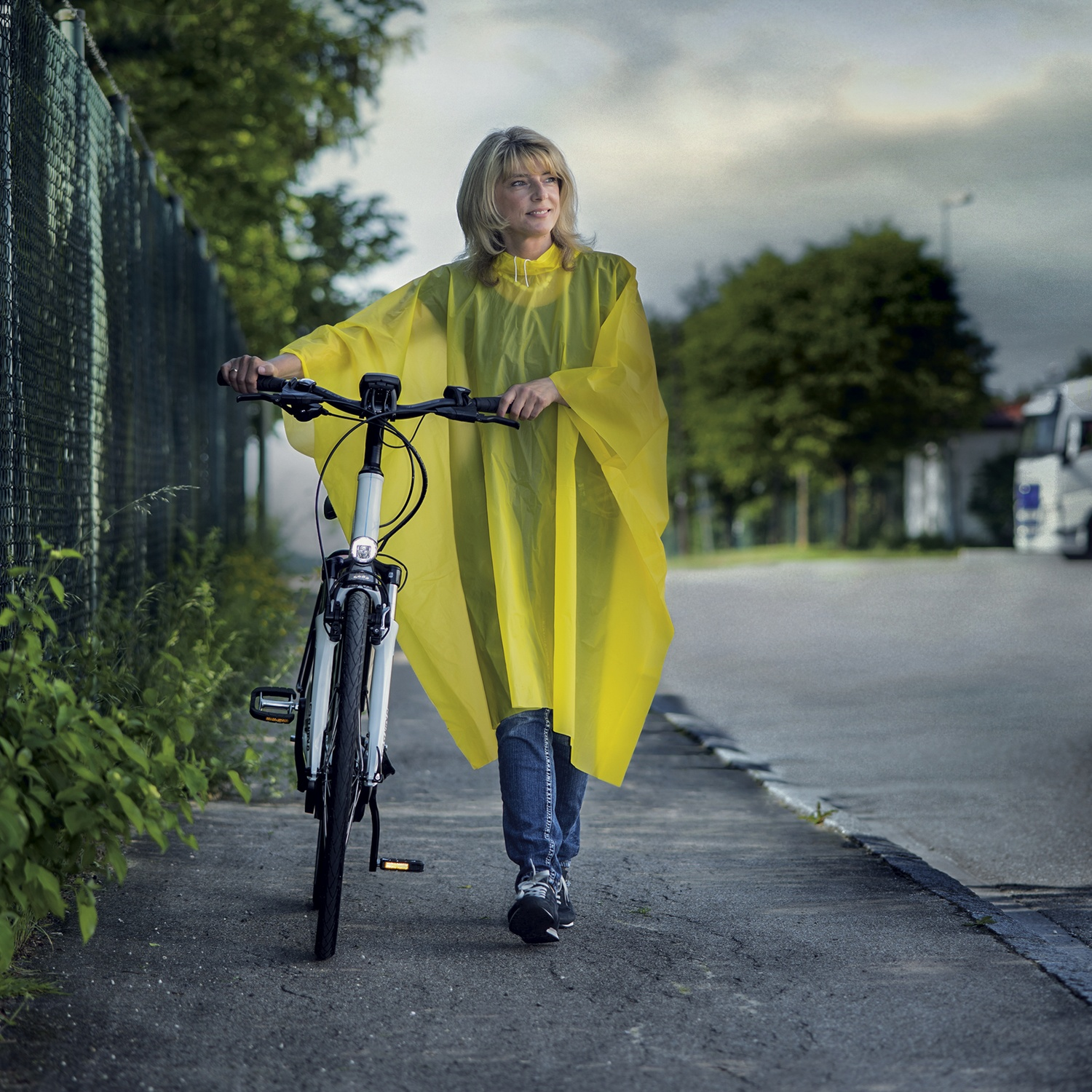 Een blonde vrouw in een gele regenponcho duwt haar fiets over een stoep.