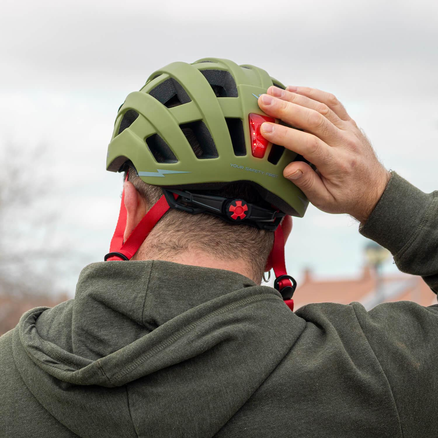 Un hombre con sudadera verde oliva se pone un casco de bicicleta verde oliva con luz trasera roja.