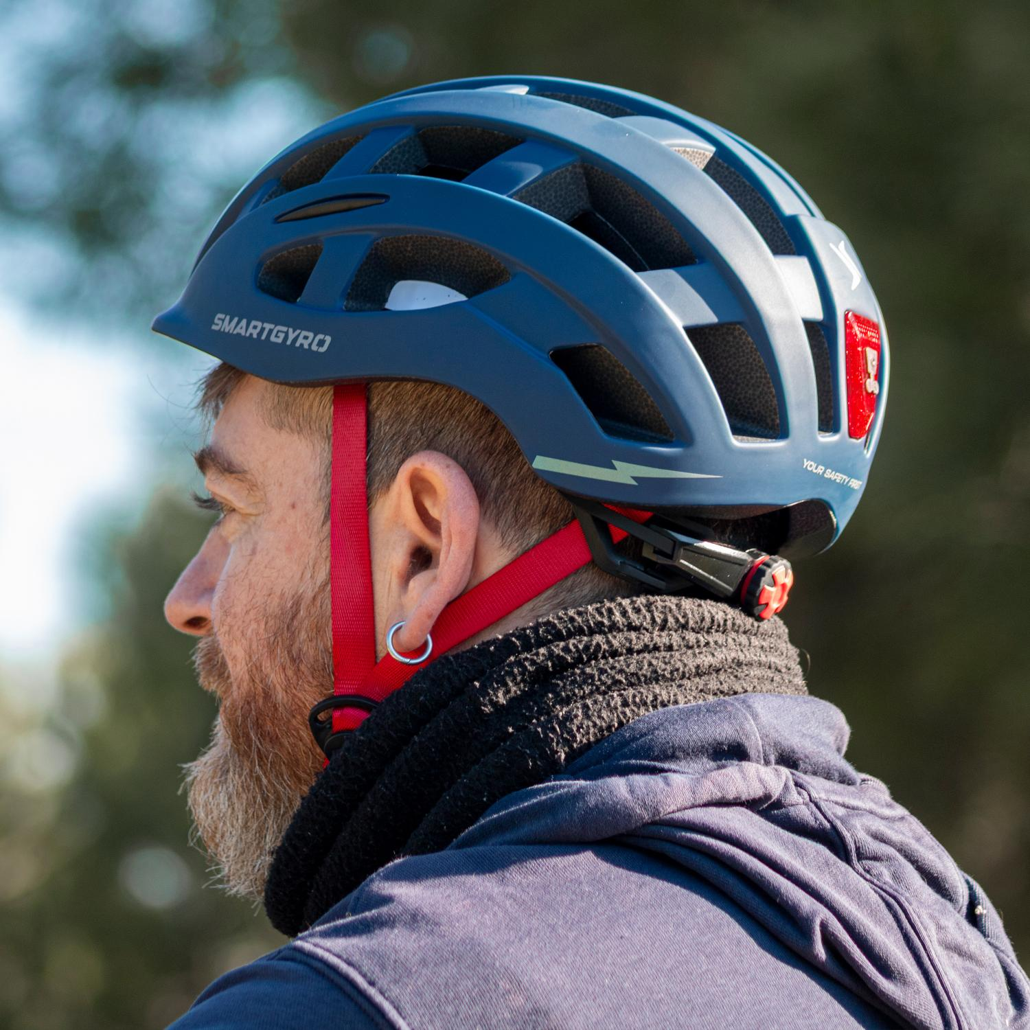 Perfil de un hombre con barba que lleva un casco de bicicleta azul, correas rojas y una sudadera con capucha oscura.