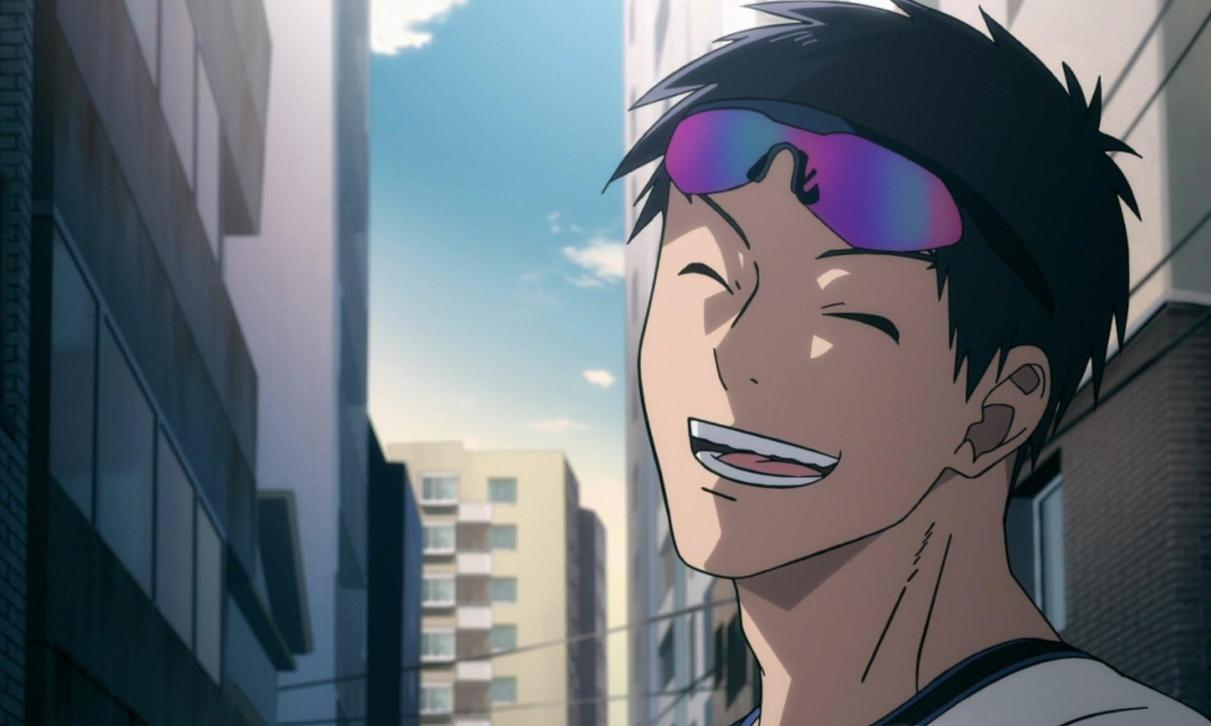 Ein lächelnder junger Mann mit schwarzen Haaren und bunter Sonnenbrille steht in einer Stadt.