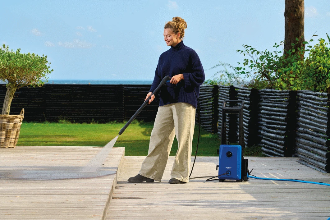 Una mujer con suéter azul limpia una terraza de madera con una hidrolimpiadora azul.