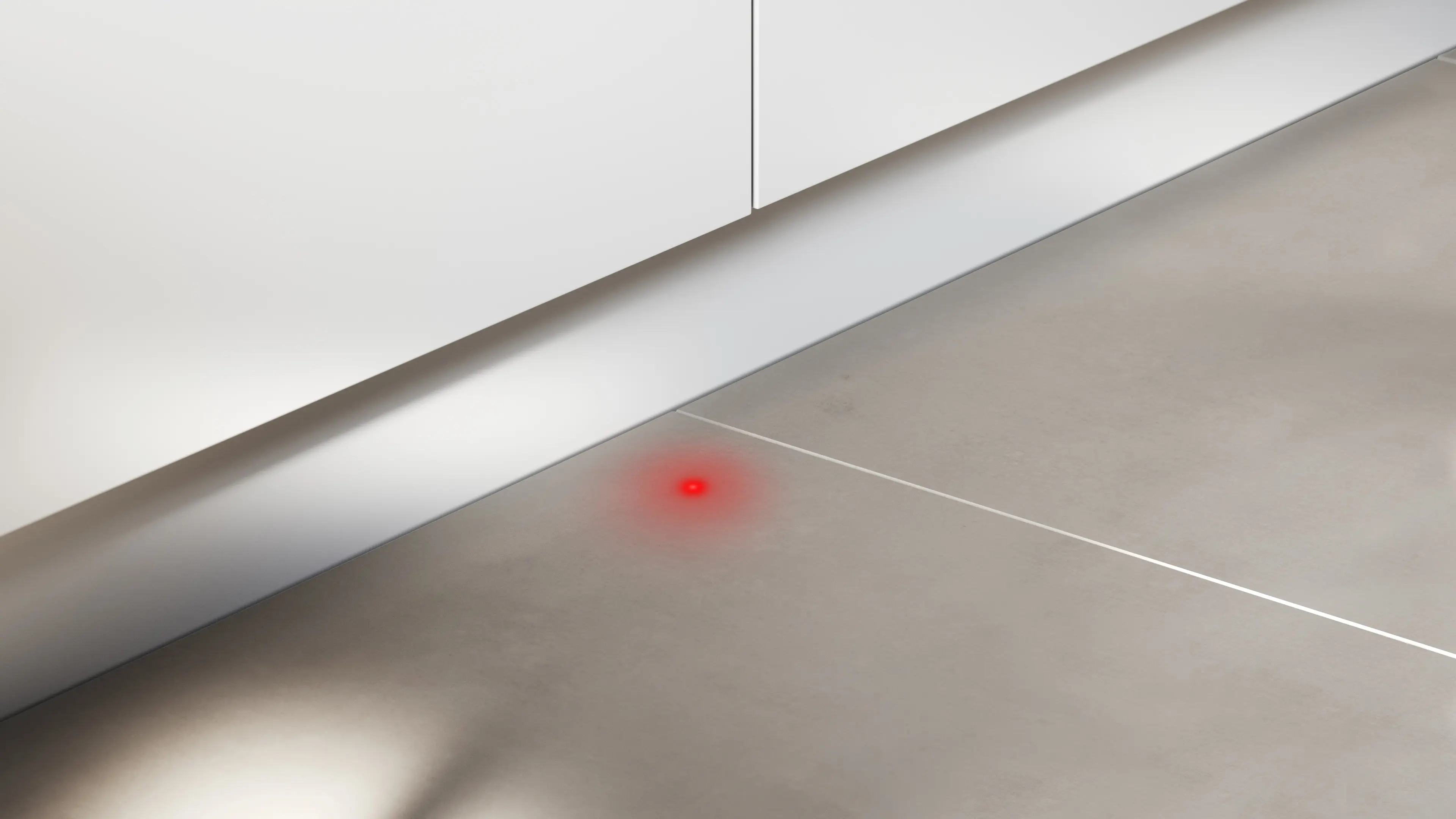 Un punto de luz roja brilla en el suelo de baldosas claras junto a los muebles de cocina blancos.