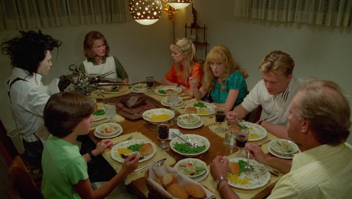 Eine Familie und Edward mit den Scherenhänden sitzen an einem reich gedeckten Esstisch unter Hängeleuchten.
