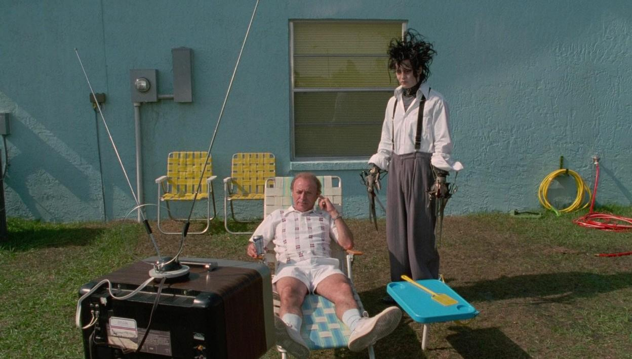 Ein Mann sitzt vor einem Fernseher auf einem Stuhl, während Edward mit Scherenhänden neben ihm steht.