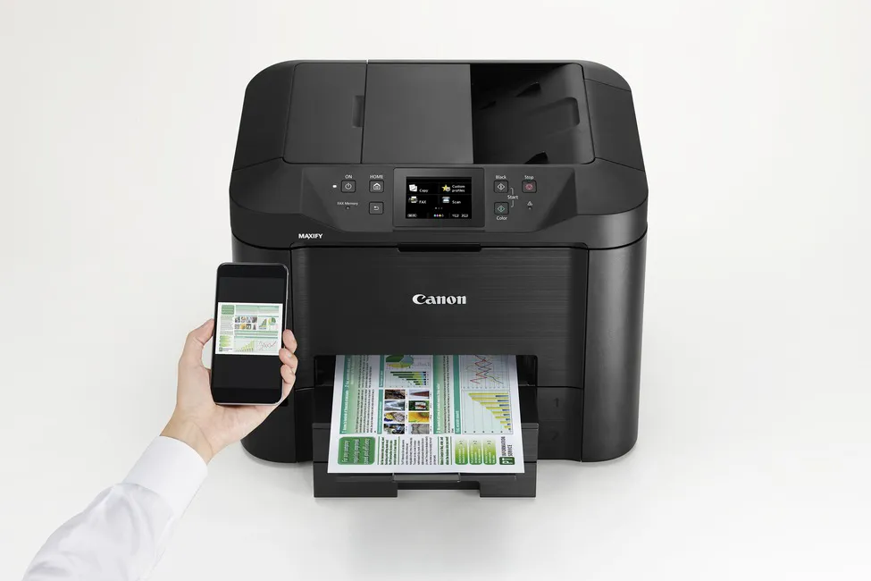 Ein schwarzer Canon Maxify Drucker druckt ein Dokument, während eine Hand ein Smartphone mit demselben Bild hält.