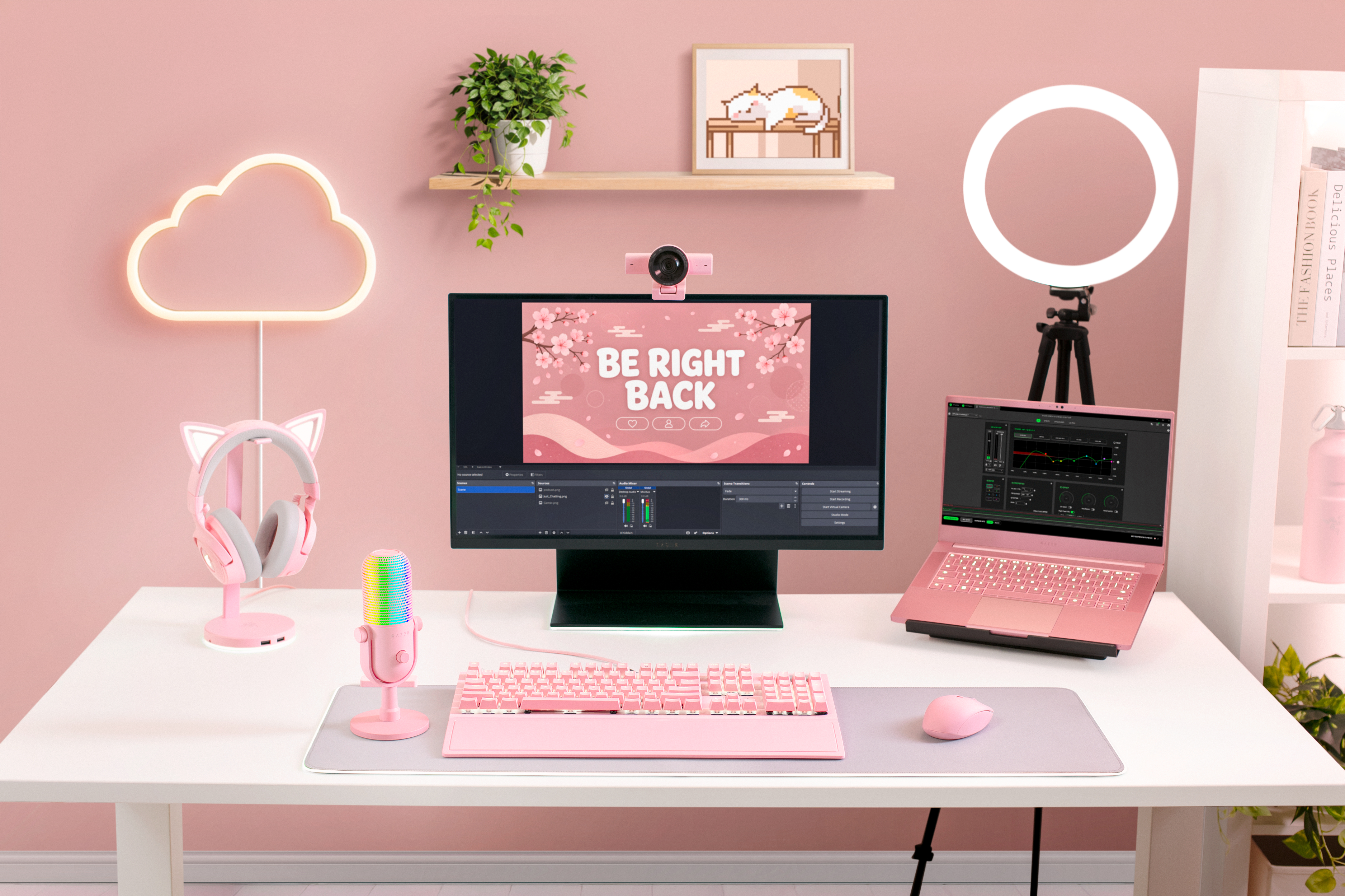 Ein rosafarbenes Gaming-Setup mit Monitor, Laptop, Mikrofon, Tastatur und einer Wolken-Neonlampe an der Wand.