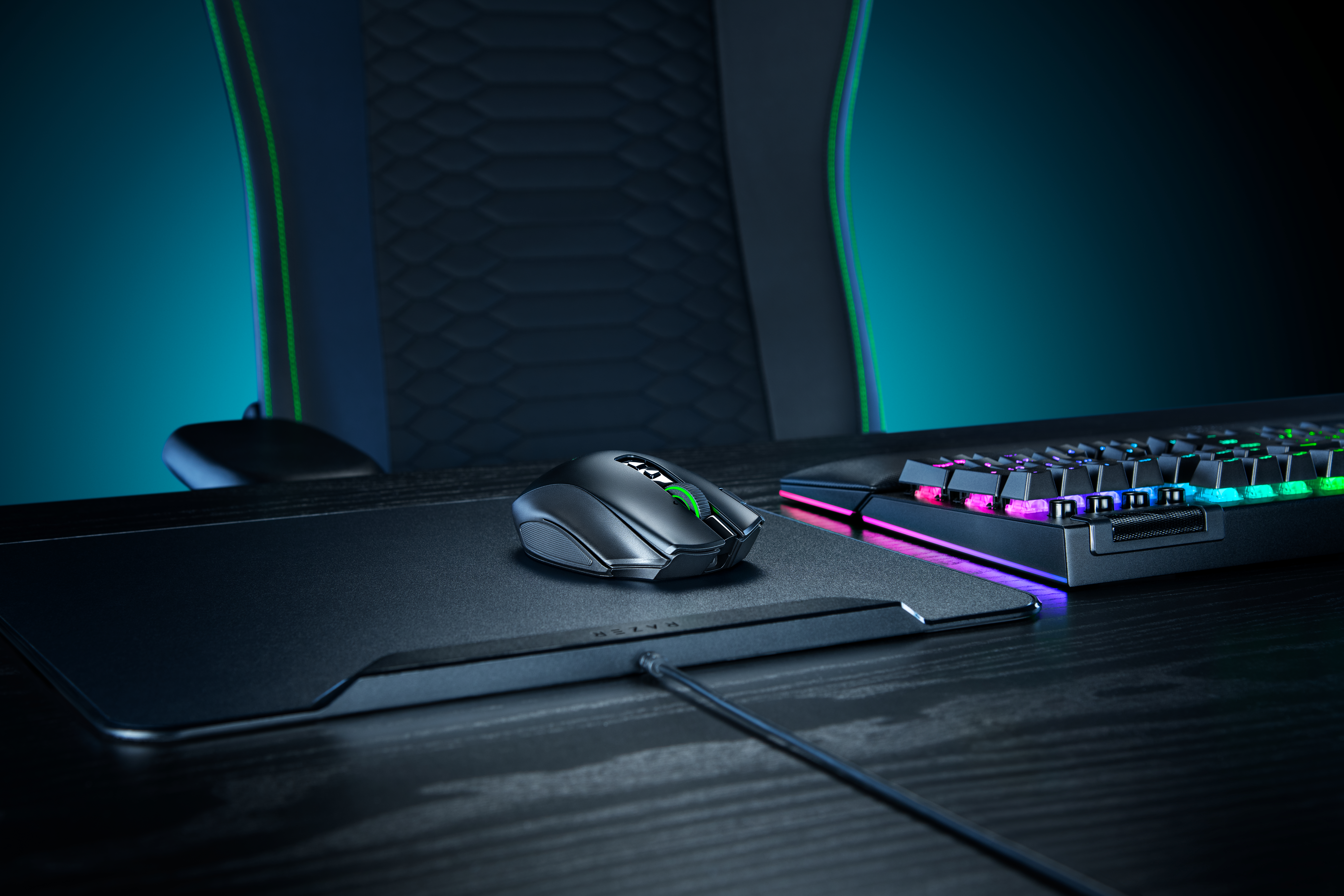 Eine schwarze Computermaus auf einem Mauspad mit RGB-Tastatur auf einem Tisch und einem Gaming-Stuhl im Hintergrund.
