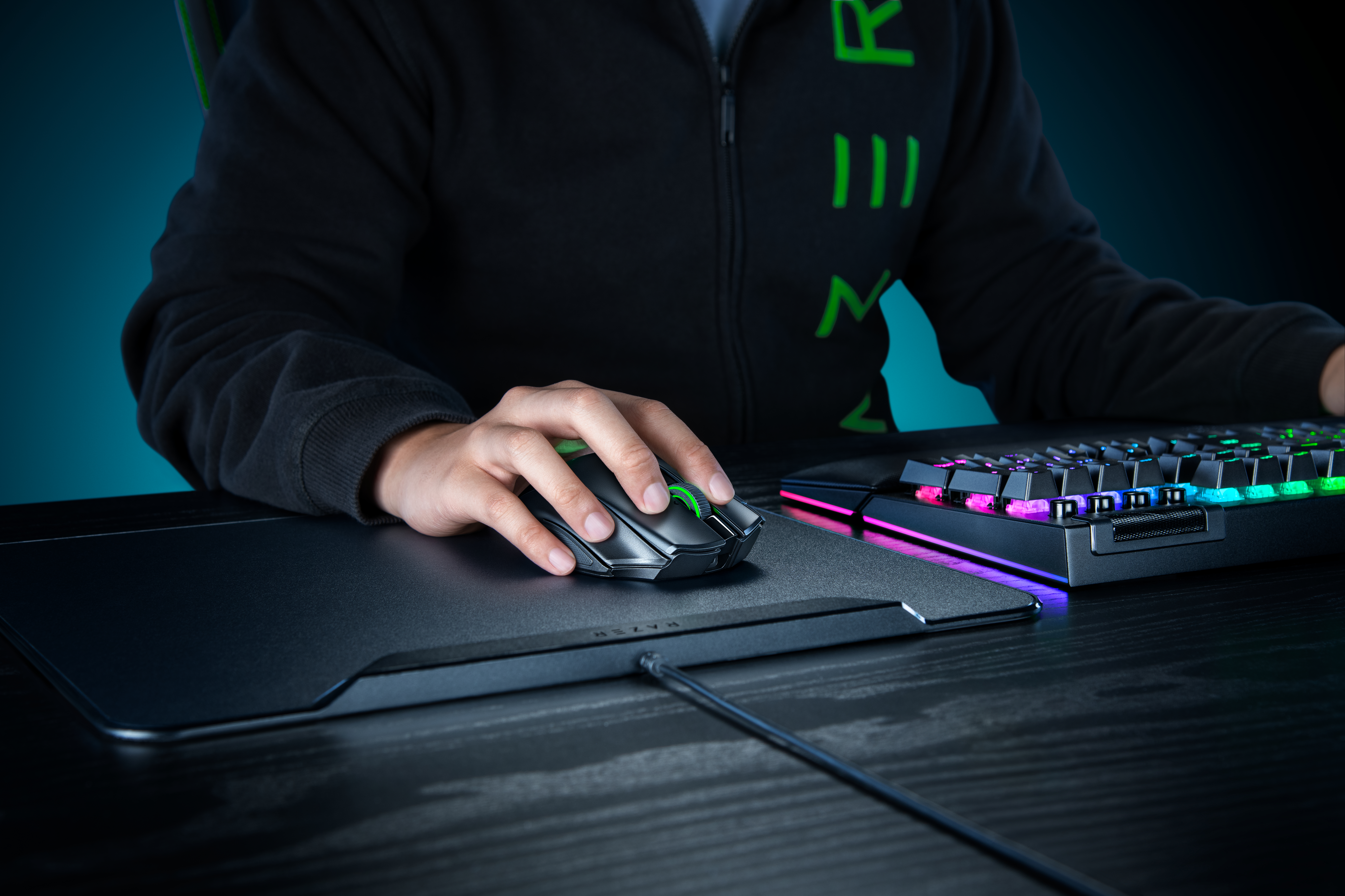 Eine Person in schwarzer Jacke nutzt eine Gaming-Maus auf einem Pad neben einer leuchtenden Tastatur.