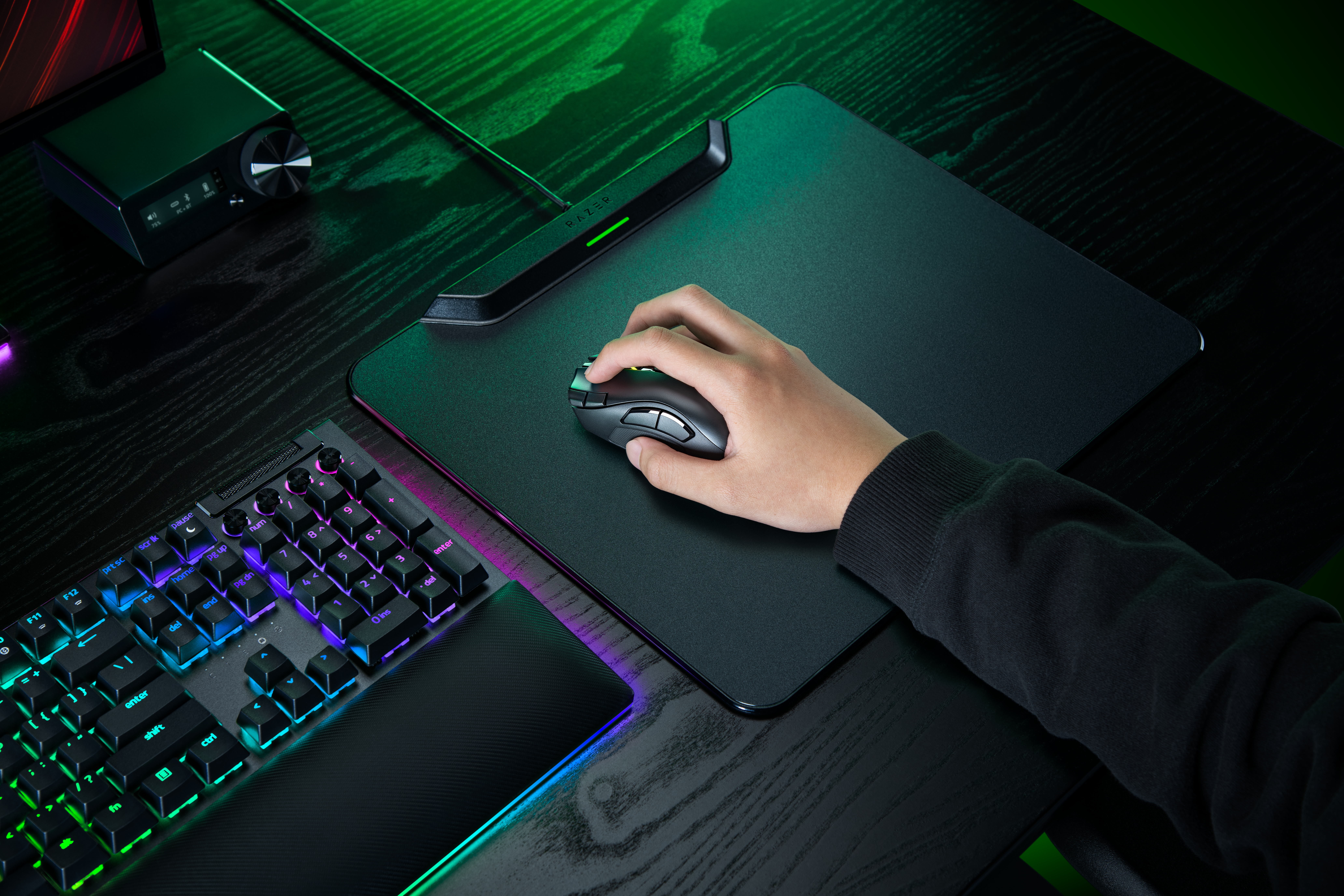 Eine Person benutzt eine schwarze Gaming-Maus auf einem Pad neben einer beleuchteten RGB-Tastatur auf einem Holzschreibtisch.