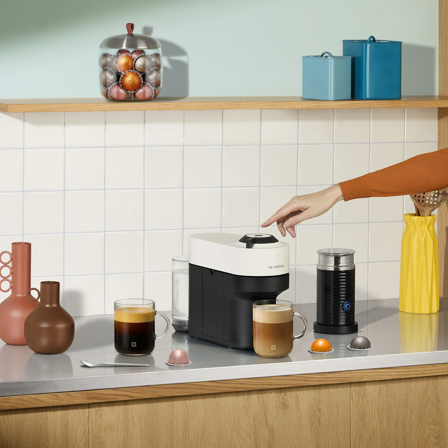 A hand points to a white and black coffee machine on a kitchen counter with mugs and capsules.