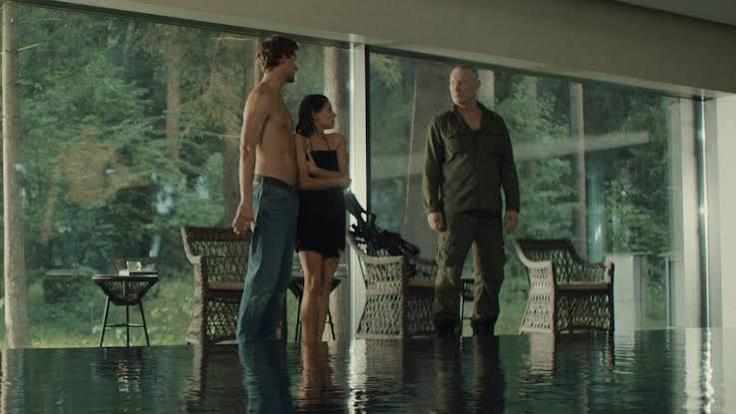 Drei Personen stehen in einem modernen Raum mit großen Glasfenstern und Blick auf einen Wald neben einem Pool.