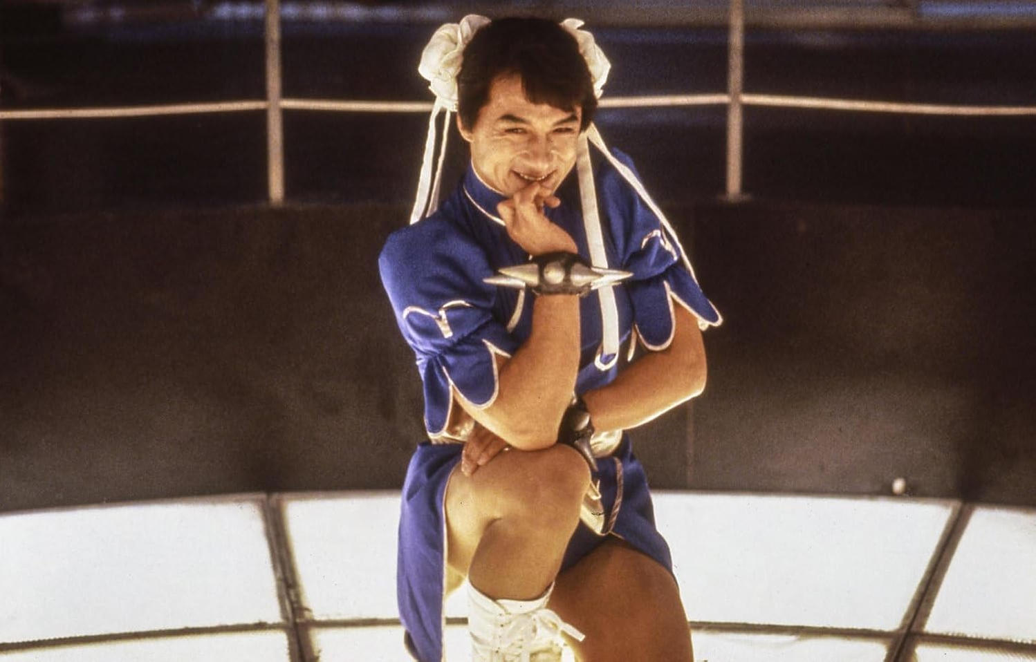 Jackie Chan kniet in einem blauen Kostüm mit weißen Schleifen und lächelt in die Kamera.