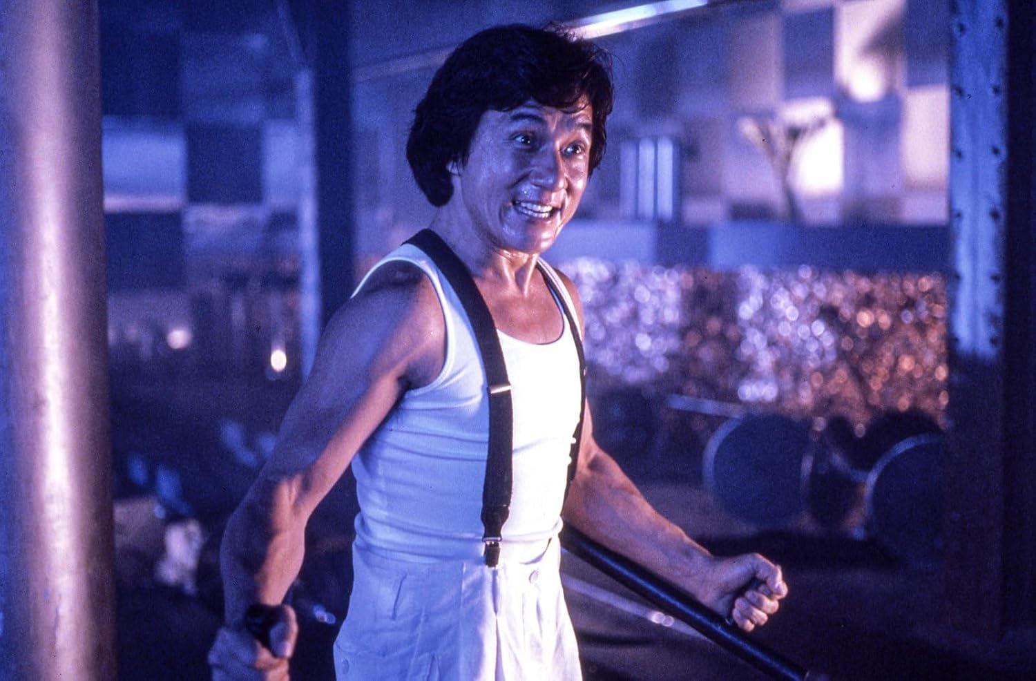 Jackie Chan in einem weißen Unterhemd und Hosenträgern lächelt und hält Metallstangen in einem dunklen Fitnessstudio.