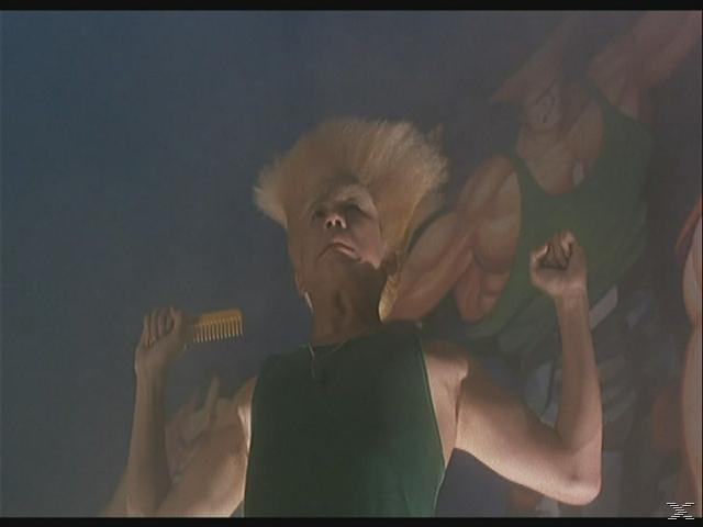 Ein Mann mit stacheligen blonden Haaren und grünem Tanktop hält einen Kamm und posiert vor einem Wandgemälde.