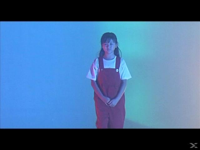 Ein Mädchen mit weißen Shirt und roter Latzhose steht vor einem blau und gelb beleuchteten Hintergrund.