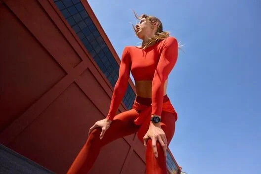 Una mujer con ropa deportiva roja posa frente a un edificio rojo bajo un cielo azul.