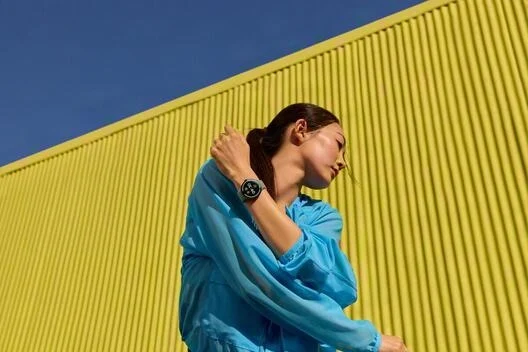 Mujer con chaqueta azul y reloj inteligente se estira ante un muro amarillo.