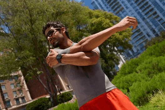 Un hombre con gafas de sol y smartwatch se estira en un parque frente a edificios.