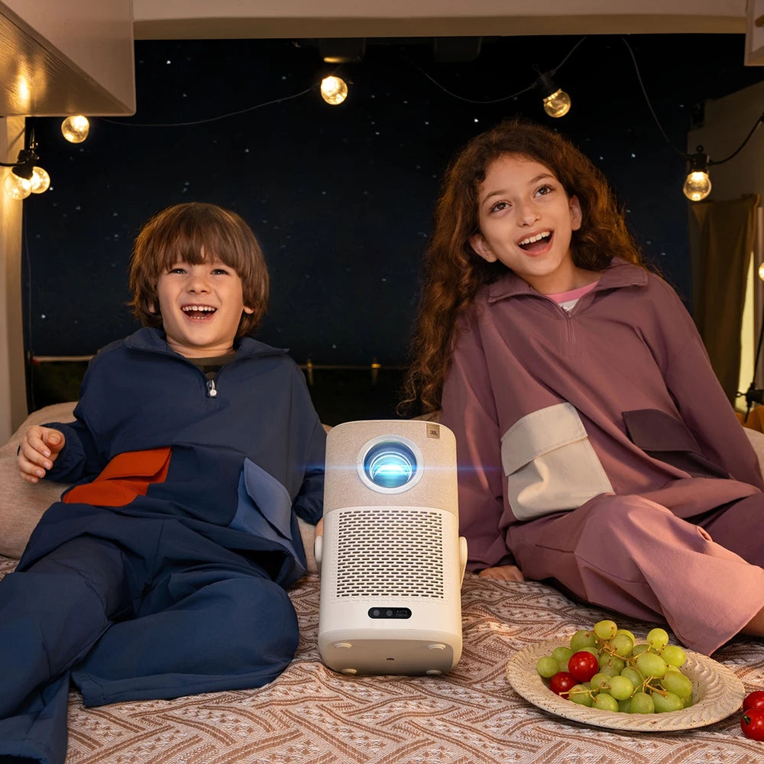 Deux enfants souriants assis près d'un projecteur sur un lit sous des guirlandes et un fond étoilé.