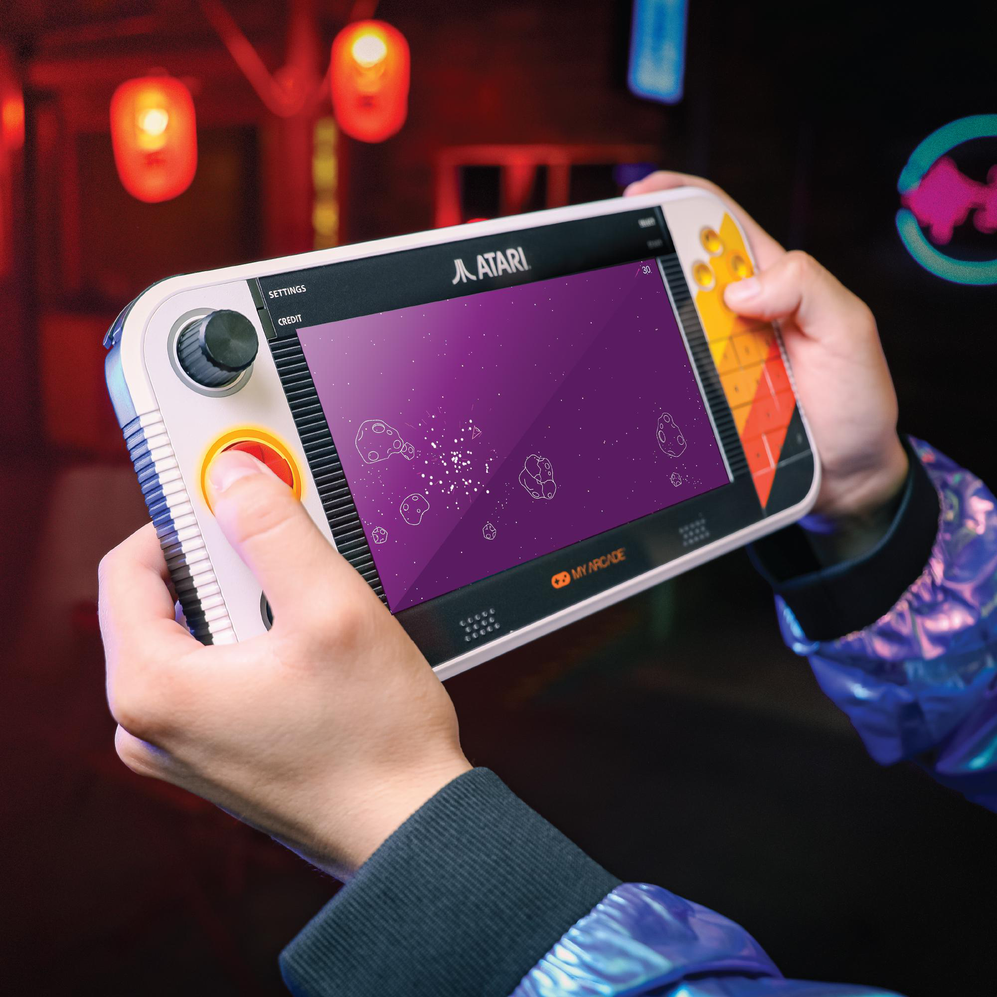 Person hält eine weiße Atari-Handheld-Konsole mit einem Weltraumspiel im Hintergrund mit Lichtern.