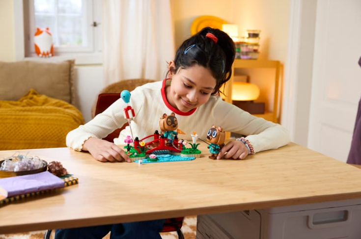 Ein lächelndes Mädchen baut Animal Crossing-Lego an einem Holztisch in einem hellen Zimmer.