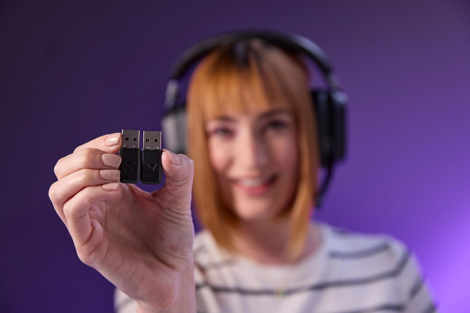 Une femme avec un casque tient deux petites clés USB noires vers la caméra sur fond violet.