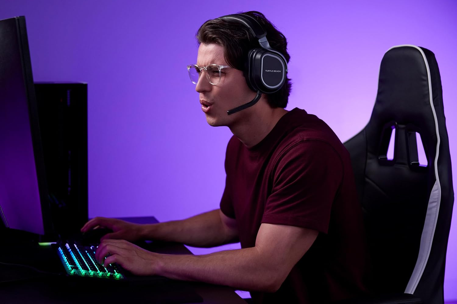 Un jeune homme avec des lunettes et un casque joue sur un ordinateur avec un clavier lumineux dans une pièce à lumière violette.