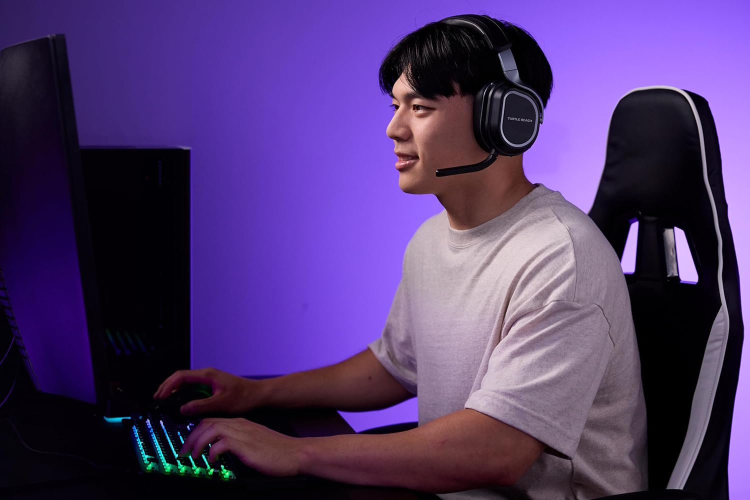 Un jeune homme portant un casque est assis à un bureau, jouant sur un ordinateur avec un clavier lumineux dans une pièce violette.