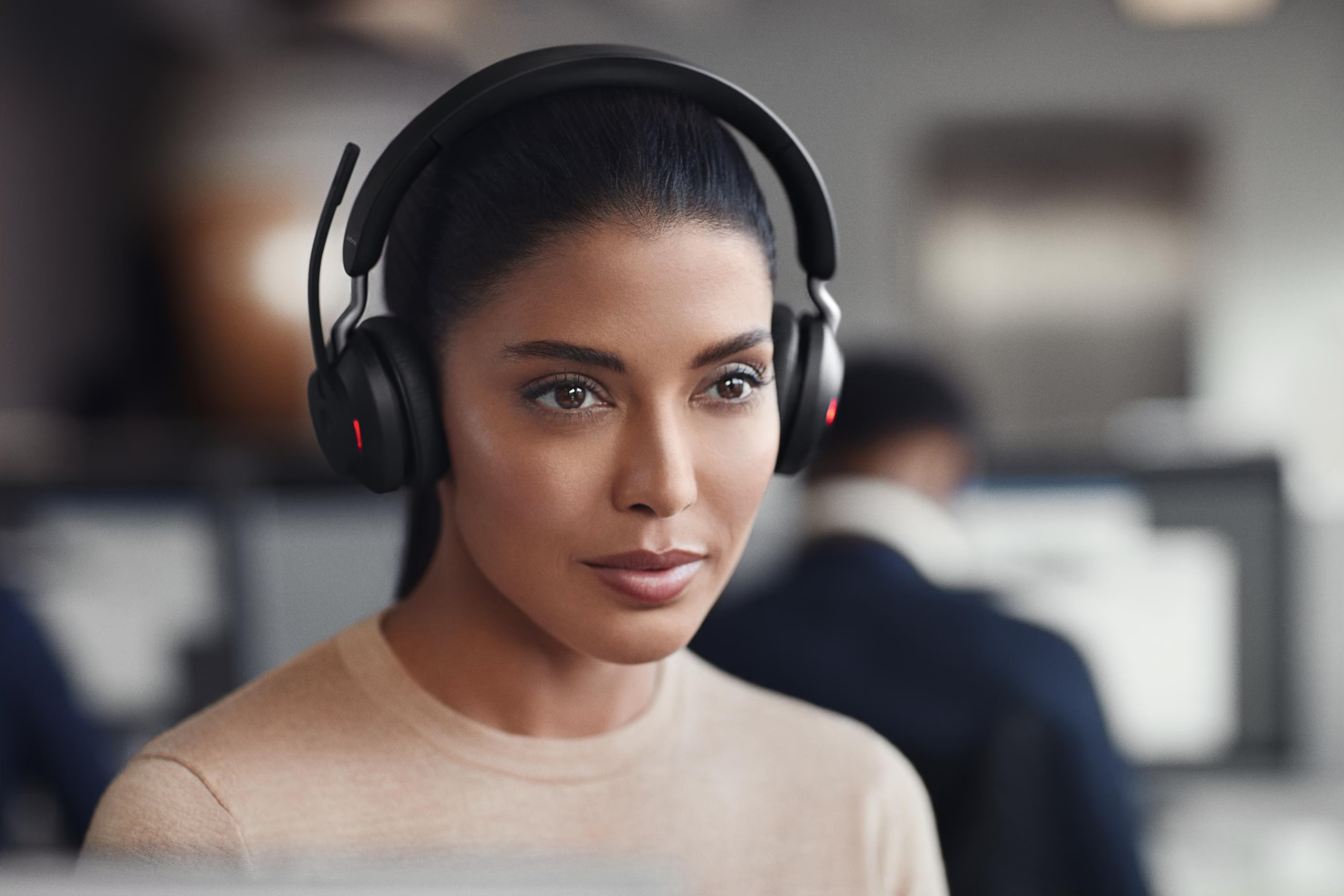Una donna con i capelli scuri indossa cuffie nere e un top beige in un ufficio.