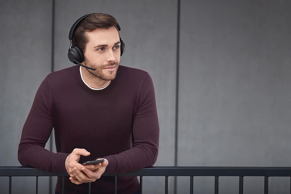 Un uomo con cuffie e maglione bordeaux si appoggia a una ringhiera tenendo uno smartphone.