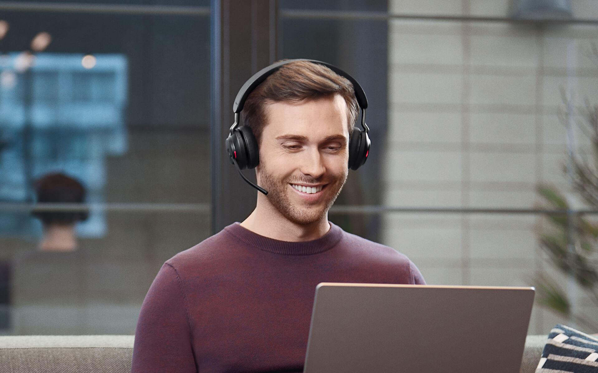Un uomo sorridente con le cuffie lavora su un computer portatile davanti a una finestra.