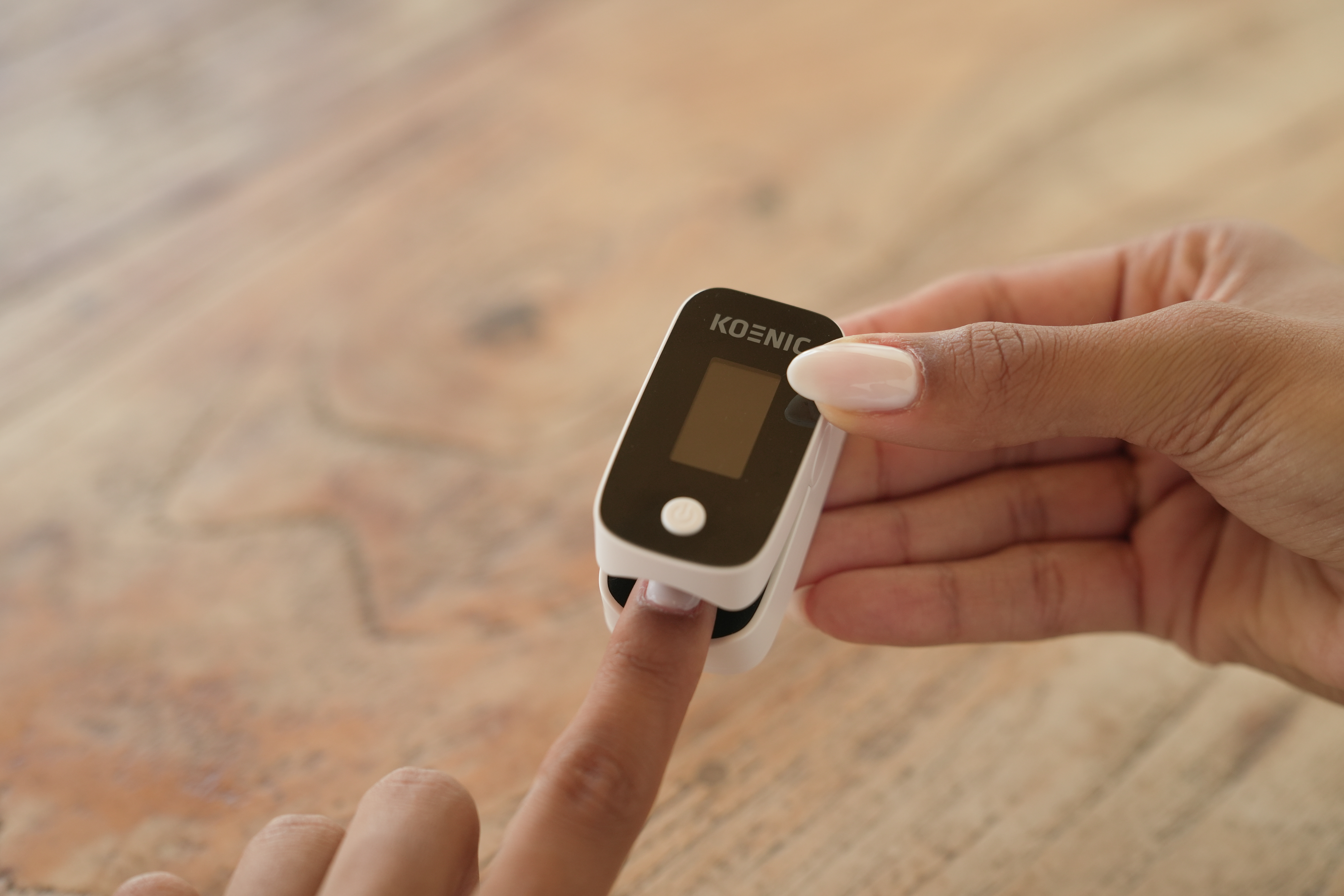 Eine Person misst den Puls mit einem weißen Pulsoximeter am Finger über einem Holztisch.