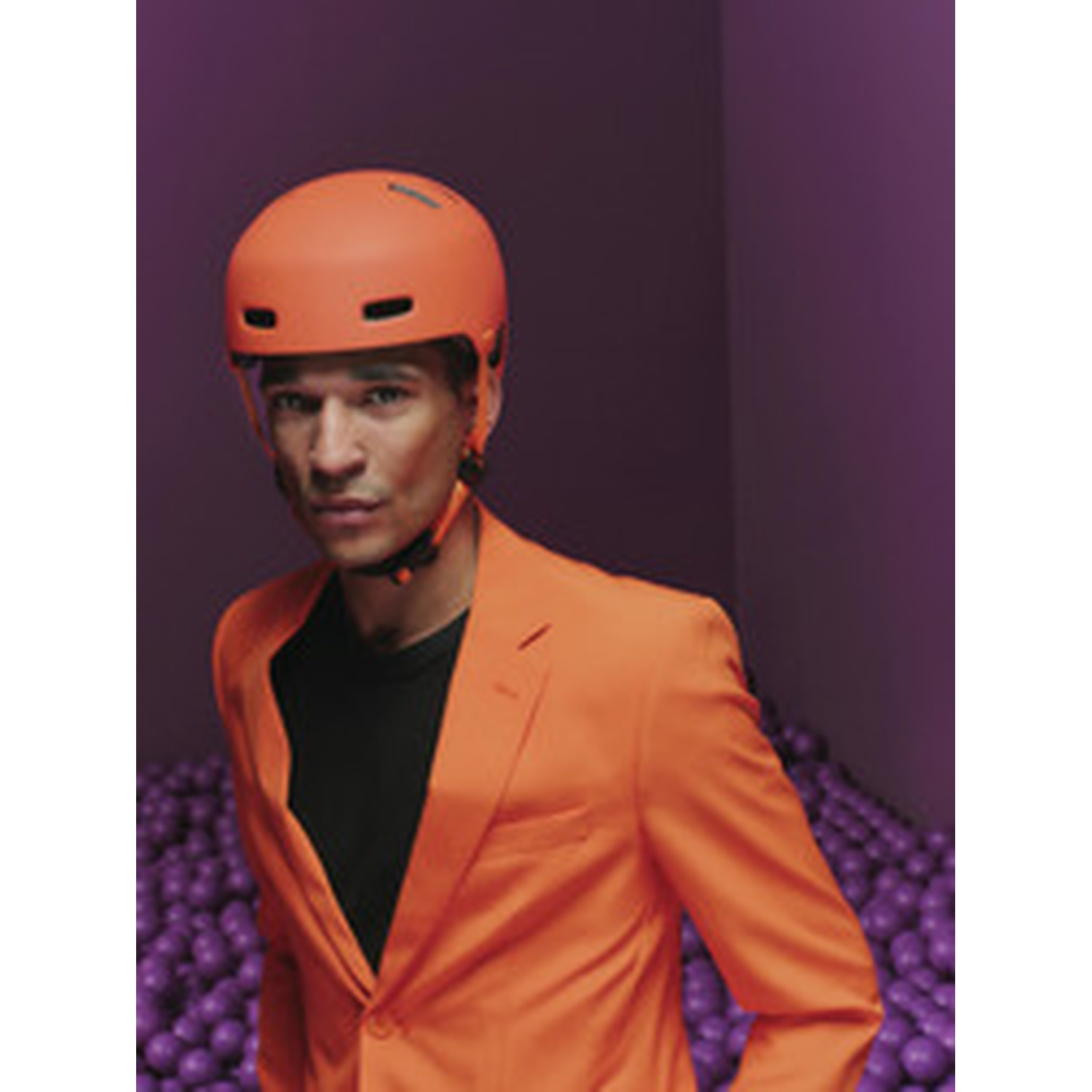 A man wearing an orange jacket and helmet stands before purple balls.