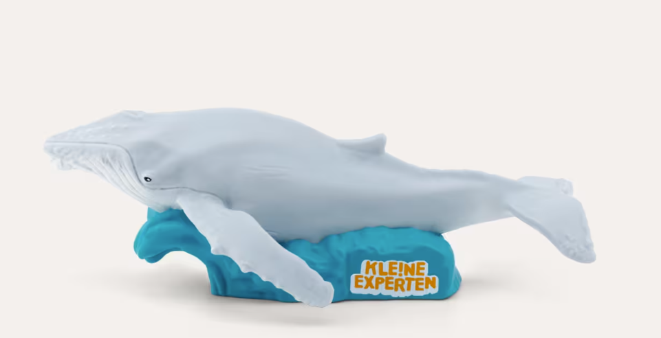 Eine graue Humpbackwal-Figur auf einem blauen Wellensockel mit dem Schriftzug KLEINE EXPERTEN auf weißem Hintergrund.