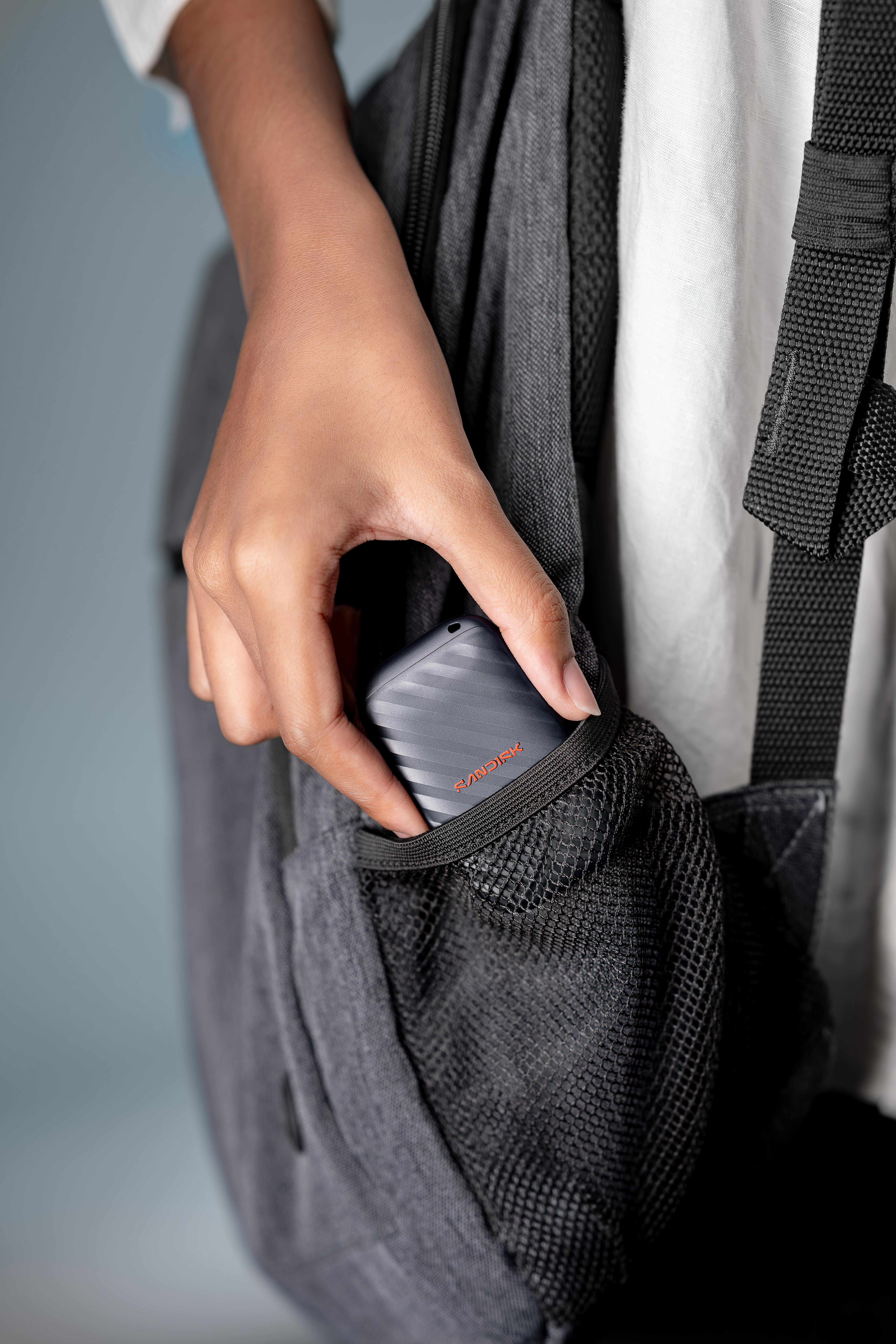 Eine Hand steckt eine kleine graue Festplatte in die Netztasche eines grauen Rucksacks.