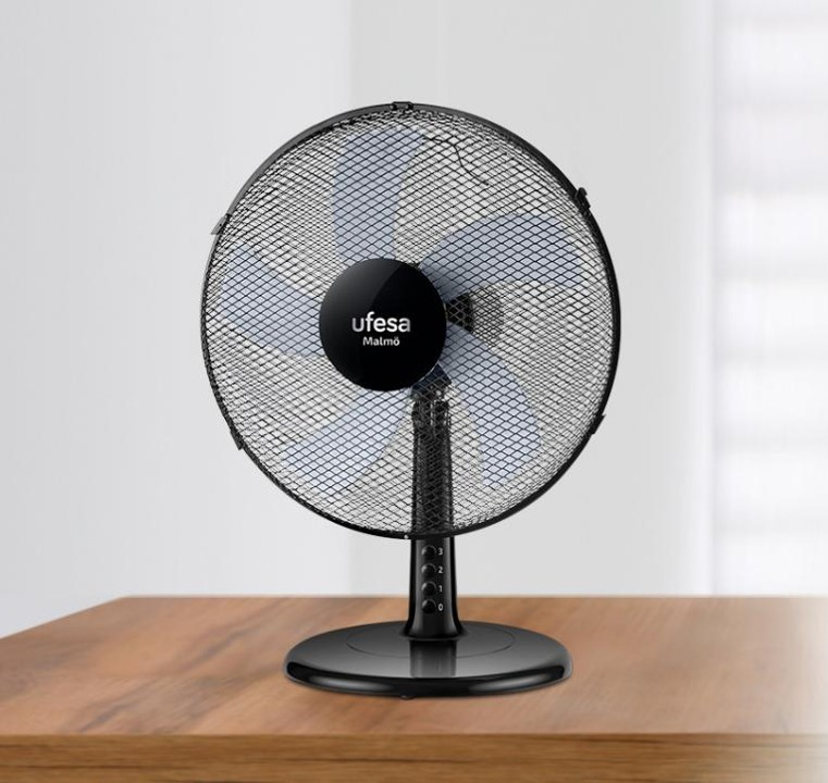 Un ventilador de mesa negro con rejilla protectora circular sobre una superficie de madera y fondo difuminado.