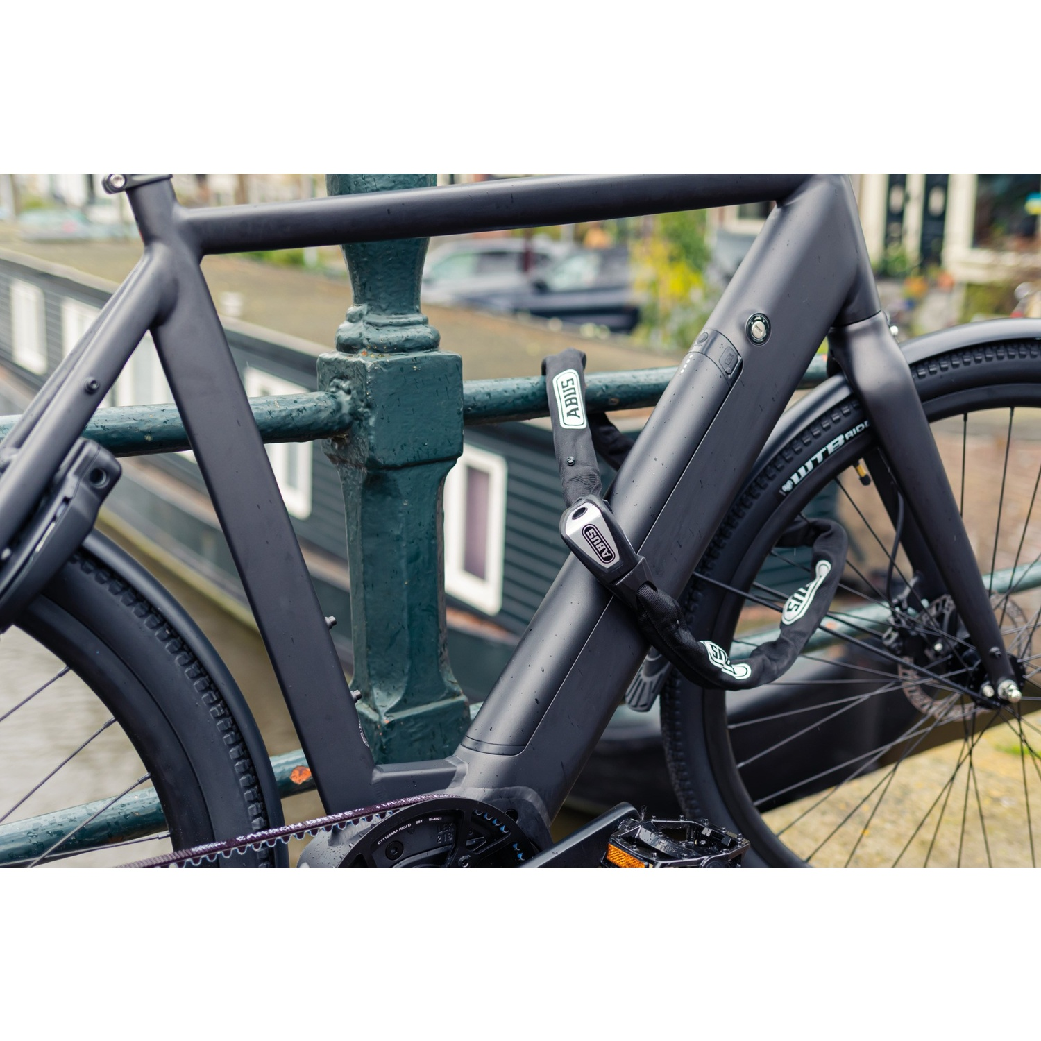 A matte black bicycle is securely locked to a green bridge railing with a black chain lock.