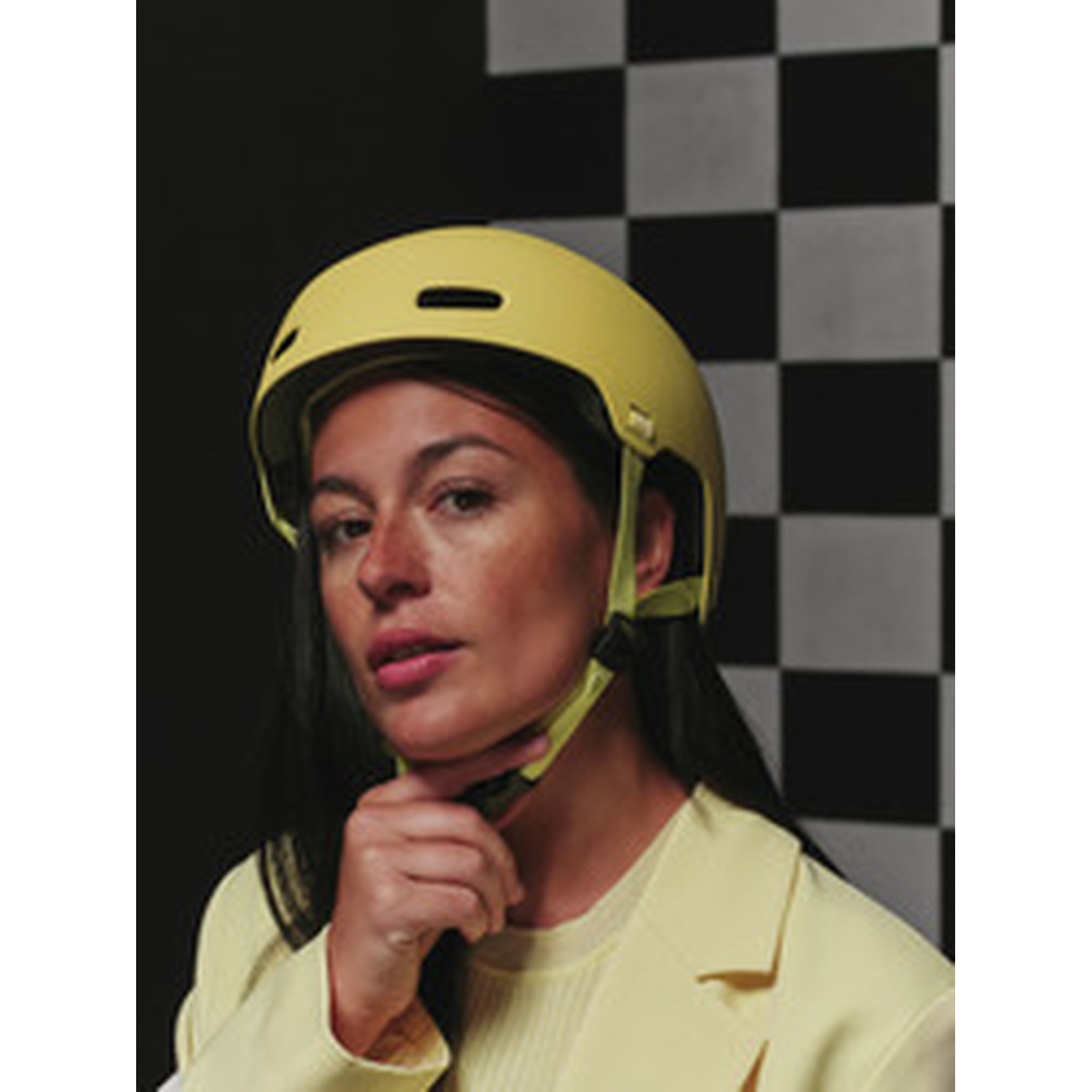 A woman in a yellow blazer and yellow helmet touches her chin strap against a black and white checkered backdrop.