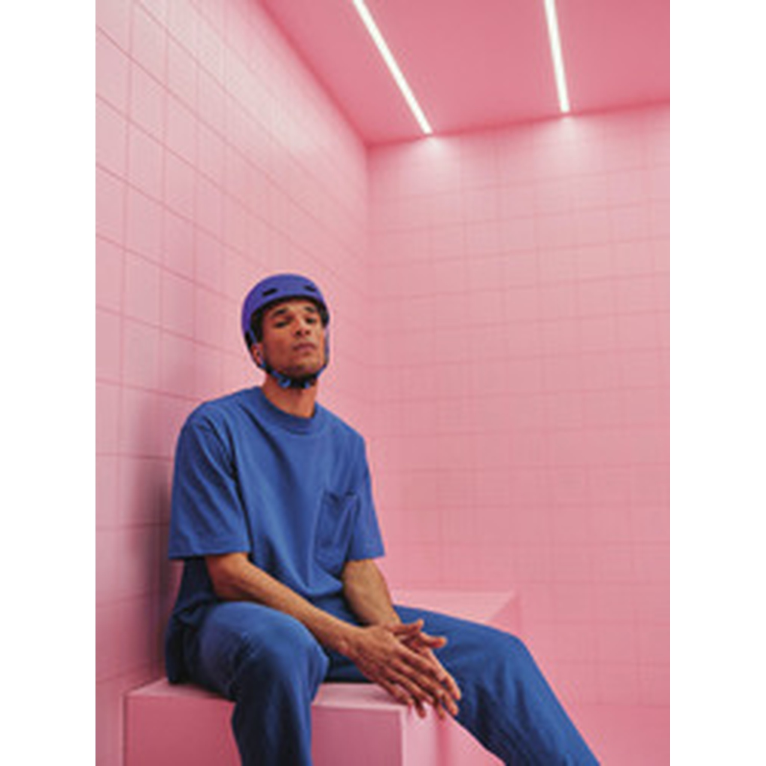 A man in a blue helmet and blue outfit sits in a pink room with tiled walls.