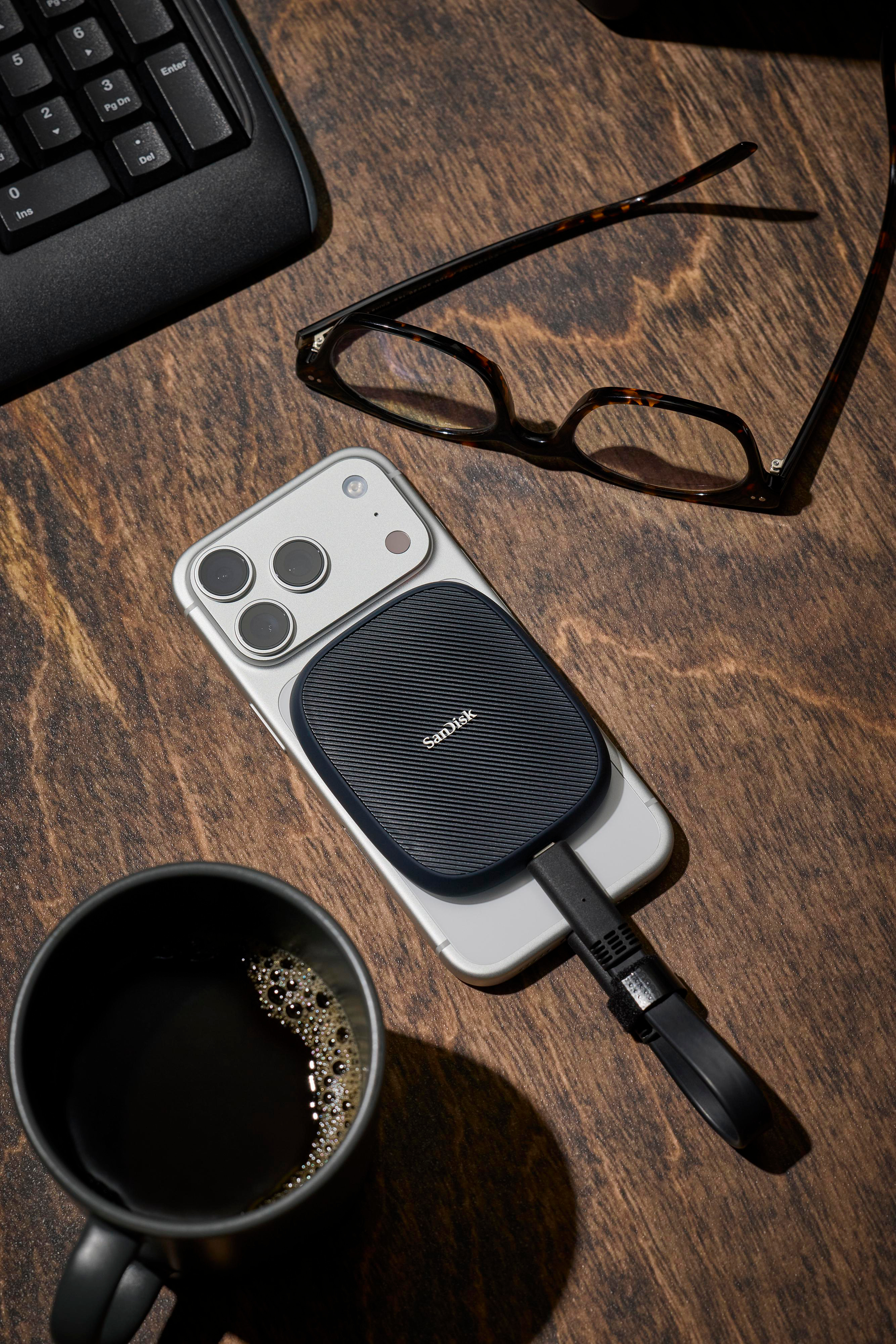 Ein Smartphone mit angeschlossener SanDisk-Festplatte, Brille, Kaffeetasse und Tastatur auf Holzschreibtisch.